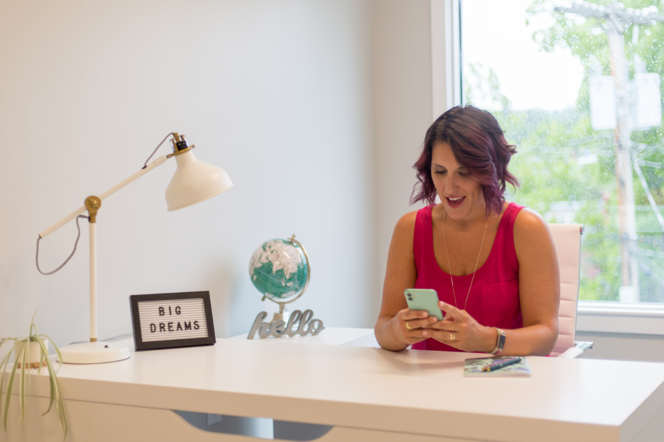 A woman at a white desk smiling at her phone, with a lamp and a small globe beside her