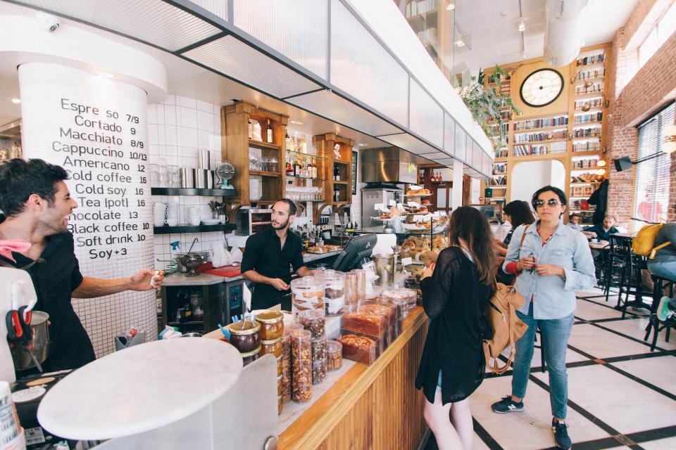 Customers ordering at the counter of a busy, bright cafe with a menu painted on a pillar