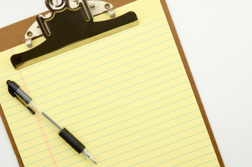 A clipboard holding a yellow legal pad with a pen resting on it